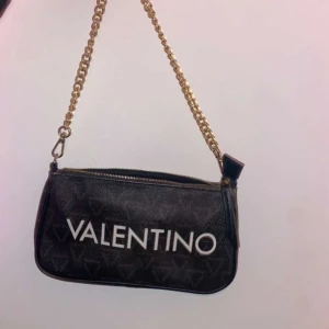 Svart axelväska från Valentino - Snygg svart axelväska från Valentino med guldfärgad kedja och dragkedja. Väskan har ett diskret mönster med Valentino-loggan och en avtagbar axelrem. Inköpt på zalando.
