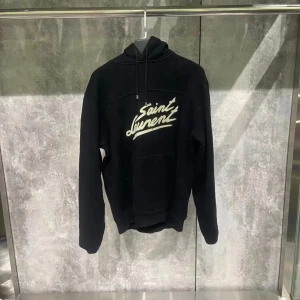 Svart hoodie från Saint Laurent - Säljer en stilren svart hoodie från Saint Laurent med vit text på framsidan. Tröjan har en klassisk design med huva och dragsko. Perfekt för en avslappnad och trendig look. Storlek S, möjligt att Pruta vid snabbköp! Mer frågor eller funderingar hör av dig!