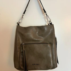 Olivgrön axelväska från Suri Frey - Snygg olivgrön axelväska från Suri Frey. Väskan har en praktisk dragkedja och dekorativa fransar. Insidan är rymlig med flera fack och ett mönstrat foder. Kommer även med ett längre väskband!