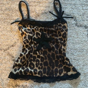 Leopardmönstrad linne med spetsdetaljer - Trendig leopardmönstrad linne med svarta spetsdetaljer och en stor stjärna framtill. Axelbanden har små rosetter och nederkanten är dekorerad med spets. 