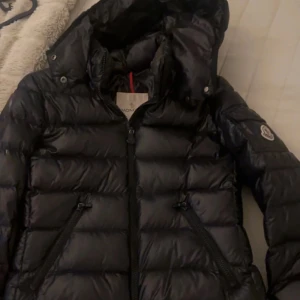 Svart dunjacka från Moncler - Snygg svart dunjacka från Moncler med huva och dragkedja. Jackan har en quiltad design och två praktiska fickor med dragkedjor. Perfekt för kyliga dagar när du vill hålla stilen. 🖤Storlek 152 :) men är större i storleken ! Den är äkta , bilder på streckkod fås privat 🤍xs-xxs
