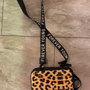 Leopardmönstrad axelväska från Forever Young - Snygg leopardmönstrad axelväska från Forever Young med justerbar axelrem. Väskan har en dragkedja för säker förvaring och är perfekt för att ge din outfit en trendig touch. 🐆