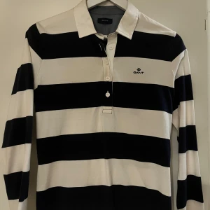 Randig pikétröja från GANT - Säljer en klassisk randig pikétröja från GANT i marinblå och vit. Perfekt för en stilren och avslappnad look.