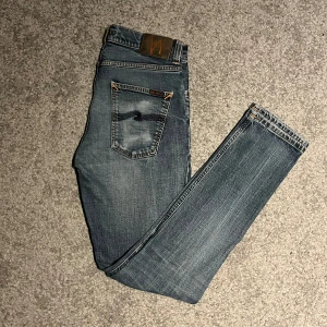 Blå jeans från Nudie - Snygga blå jeans från Nudie med klassisk femficksdesign och en diskret tvätt för en avslappnad look. De har en dragkedja och knapp framtill samt märkets logga på bakfickan. Perfekta för en stilren vardagsstil.