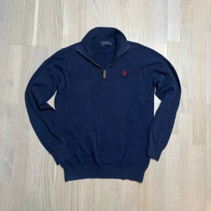 Mörkblå tröja med dragkedja från Polo Ralph Lauren - Säljer en stilren mörkblå tröja från Polo Ralph Lauren med dragkedja och röd broderad logotyp på bröstet. Tröjan har långa ärmar och en ribbad krage. Perfekt för en avslappnad och snygg look.