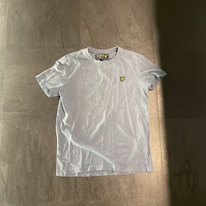 Ljusblå tröja från Lyle & Scott Junior - Säljer en stilren ljusblå t-shirt från Lyle & Scott Junior med det klassiska gula logotypen på bröstet. T-shirten är kortärmad och tillverkad i mjukt bomullsmaterial. Perfekt för en avslappnad look. Köpt på Lyle & Scott.