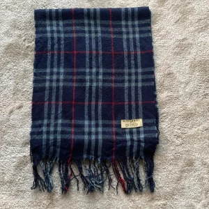 Rutig halsduk från Burberry - Snygg rutig halsduk från Burberry i mörkblått med röda och ljusblå ränder. Tillverkad i 100% lammull och har fransar i ändarna. Perfekt för att hålla dig varm och stilren under kyliga dagar. Pris 449 kronor, inte hugget i sten 