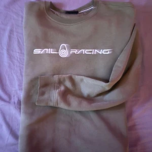 Olivgrön tröja från Sail Racing - Säljer en olivgrön tröja från Sail Racing med deras logga tryckt i vitt på bröstet. Tröjan har en klassisk rund hals och långa ärmar, perfekt för en avslappnad stil. Passar bra till både jeans och chinos. Är i ( L) men passar både ( L & M )