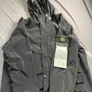 Svart jacka från Stone Island - Säljer en snygg svart jacka från Stone Island. Jackan är tillverkad i polyester med en vind- och vattenavvisande yta. Den har en huva och med dragkedja och knappar framtill. Hör av er om ni har frågor! Nypris 6500 men har inte kommit til användning!