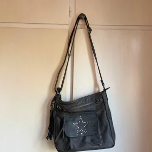 Svart axelväska med stjärndetalj - Säljer en snygg ”smuts” grå  axelväska i skinn med en cool stjärndetalj på framsidan. Väskan har en dragkedja upptill och en justerbar axelrem. Perfekt för både vardag och fest!