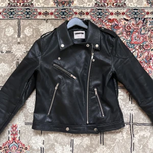 Svart skinnjacka med dragkedjor - Säljer en snygg svart skinnjacka med coola dragkedjedetaljer. Jackan har en klassisk bikerstil med asymmetrisk dragkedja framtill och flera fickor med dragkedjor. Perfekt för höst och vår, och ger en edgy look till vilken outfit som helst. Passar både till vardags och fest!