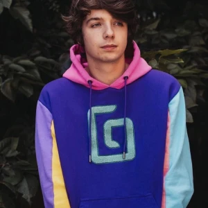 Färgglad hoodie från Jschlatt - Säljer en supercool och färgglad hoodie från Karl Jacobs. Hoddien har lila bas med en stor turkos logga på framsidan. Ärmarna är i olika färger: en är gul och lila, och den andra är turkos och rosa. Huvan är rosa med lila dragsnören. Ren och hel☀️