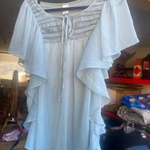 Ljusblå klänning från Vila - Säljer en superfin ljusblå blus från Vila i storlek M. Den har vackra spetsdetaljer på bröstet och en söt knytning framtill. Blusen har korta ärmar med volanger som ger en romantisk känsla. Perfekt för sommarens alla fester!