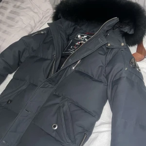 Svart dunjacka från Moose Knuckles - Säljer en snygg och varm svart dunjacka från Moose Knuckles. Jackan har en fluffig pälsfodrad huva och är perfekt för kalla vinterdagar. Den har dragkedja och flera praktiska fickor. Märket är känt för sin kvalitet och stil. Perfekt för dig som vill hålla dig varm och se bra ut samtidigt!