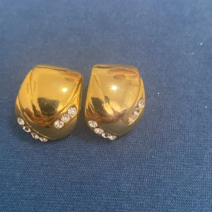 Guldiga örhängen med strassdetaljer - Säljer ett par snygga guldiga örhängen med glittrande strassdetaljer. De har en elegant och tidlös design som passar perfekt för festliga tillfällen. Örhängena är clips, vilket gör dem bekväma att bära även om du inte har hål i öronen.