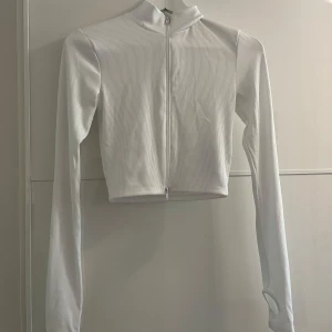 Vit ribbad croppad tröja från New Yorker  - Säljer en superfin vit ribbad tröja från New Yorker. Den är croppad och har en dragkedja framtill. Tröjan är långärmad och har hål för tummarna, vilket ger en cool och bekväm stil. Perfekt för både vardag och fest!