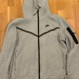 Nike tech flees - Säljer en grå huvtröja från Nike med dragkedja och svart detaljer. Tröjan har en justerbar huva och en ficka på ärmen. Perfekt för en sportig look eller avslappnade dagar.