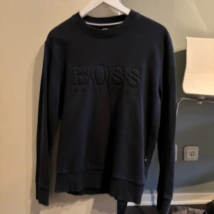 Svart sweatshirt från Boss - Säljer en stilren svart sweatshirt från Boss med broderad logga på framsidan. Tröjan har långa ärmar och en rund halsringning. Perfekt för en avslappnad look.