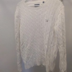 Vit stickad tröja från GANT - Säljer en stilren vit stickad tröja från GANT i premium bomull. Tröjan har ett klassiskt kabelstickat mönster och rund halsringning. Perfekt för en elegant och avslappnad look.