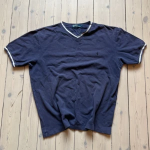 Ralph Lauren Mörkblå tröja  - Säljer en klassisk mörkblå pikétröja från Ralph Lauren med v-ringning och vita detaljer vid krage och ärmslut. Perfekt för en stilren look. Tröjan har korta ärmar och är i en bekväm passform.