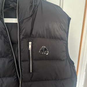 Moose knuckles väst herr  - Snygg svart dunväst med dragkedja och en cool logga på bröstet. Västen har en hög krage och praktiska fickor med dragkedjor. Perfekt för att hålla värmen under kyliga dagar. Storlek XXL sitter som XL 