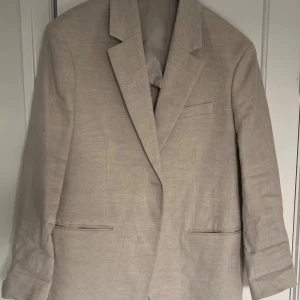 Beige linnekavaj från Arket - Snygg beige linnekavaj från Arket i en klassisk design. Perfekt för en stilren look. Kavajen har en enkel knäppning och två fickor framtill. Passar utmärkt till både formella och avslappnade tillfällen.