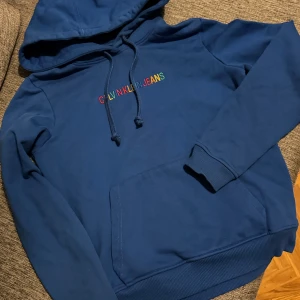 Blå hoodie från Calvin Klein Jeans - Säljer en snygg blå hoodie från Calvin Klein Jeans med färgglad logga på bröstet. Tröjan har en klassisk design med huva och dragsko samt ribbade muddar vid ärmslut och nederkant. Perfekt för en avslappnad stil.