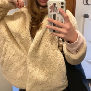 Beige teddyjacka med huva - Mysig beige teddyjacka från Zara med huva. Jackan har en fluffig och varm design, perfekt för kyligare dagar. Den har en enkel stil utan synliga knappar eller dragkedja, vilket ger en avslappnad look.