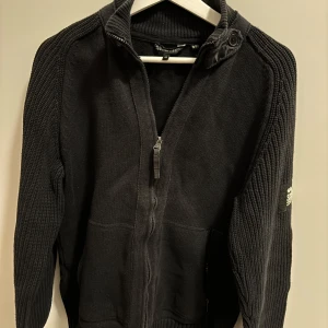 Svart stickad tröja från Crosshatch - Säljer en svart stickad tröja från Crosshatch Black Label. Tröjan har en dragkedja framtill och långa ärmar med ribbade detaljer. Perfekt för kyligare dagar. Nypris 800