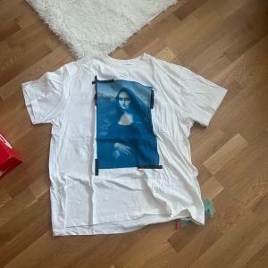 Vit t-shirt med Mona Lisa-tryck - Säljer en vit t-shirt från Off-White med ett stort Mona Lisa-tryck på framsidan och ikoniska pilar på baksidan. Perfekt för dig som vill ha en unik och konstnärlig stil. T-shirten har en normal passform och korta ärmar.