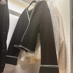 Slutsåld gina kappa - Xs men passar s Snygg svart omlottkofta från Gina med vita kontrastsömmar. Koftan har långa ärmar och en stilren design som passar perfekt för kyligare dagar. Perfekt för dig som vill ha något både bekvämt och trendigt.