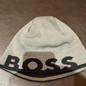 Svart och grå mössa från Boss - Mössa från Boss i svart och grått. Mössan har en stor logga i svart, medan den andra sidan har en diskret logga och en grå rand. Perfekta för kyliga dagar med en modern touch. Den är även tvåsidig.