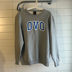 Grå sweatshirt från OVO - Säljer en grå sweatshirt från OVO med blå och vit text på framsidan. Tröjan har långa ärmar och en liten uggla broderad på ärmen. Perfekt för en avslappnad stil. På tröjan står de att strl är extra small men skulle säga att den sitter mer som en Vanlig small.