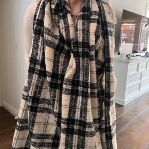 Beige och svart rutig halsduk - Snygg och trendig halsduk i beige och svart med rutigt mönster. Perfekt för att hålla dig varm och stilren under kyliga dagar. Den är stor och kan bäras på flera olika sätt för att passa din stil.