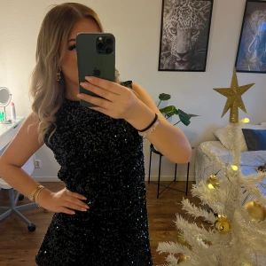 Svart glittrig klänning med rosett - Snygg svart glittrig klänning med en stor vit rosett i ryggen. Perfekt för festliga tillfällen. Klänningen är ärmlös och har en kort längd som ger en elegant look.