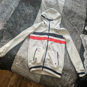 Vit hoodie från Race Marine - Säljer en stilren vit hoodie från Race Marine med dragkedja och huva. Den har ett sportigt utseende med ränder i marinblått och rött över bröstet. Perfekt för en avslappnad stil. 🧢 