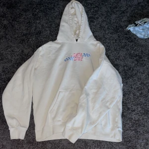 Vit hoodie från Santa Cruz - Säljer en vit hoodie från Santa Cruz med tryck i rosa och blått på både fram- och baksidan. Den har en klassisk känguruficka och en bekväm huva. Perfekt för en avslappnad stil! OBS: ACCEPTERA INTE TILLBAKA SKICKNINGAR 