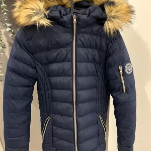 Mörkblå dunjacka med pälsdetaljer - Snygg mörkblå dunjacka med fuskpäls på huvan och dragkedja framtill. Jackan har en quiltad design och en rund logotyp på ärmen. Perfekt för kyliga dagar. Barn storlek 14 🟰 med 36