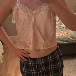 Rosa satinlinne med spetsdetaljer - Säljer ett sött rosa linne i satin med smala axelband och vacker spetskant. Perfekt för en somrig look eller som ett lager under en tröja. Linnet har en lös passform och är croppad för en modern stil.