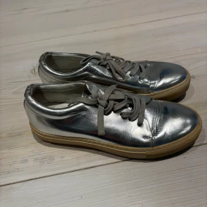 Silvriga sneakers från Acne Studios - Snygga silvriga sneakers från Acne Studios med snörning och en klassisk design. Skorna har en glansig yta och en bekväm sula i gummi. Perfekta för att ge din outfit en modern touch.
