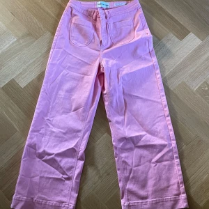 Rosa högmidjade byxor från Tomorrow - Snygga rosa byxor från Tomorrow med hög midja och flare-stil. Byxorna har framfickor och en dragkedja. Perfekta för en färgglad look!