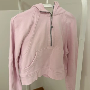 Ljusrosa hoodie med dragkedja - Säljer en ljusrosa hoodie från Lululemon med halv dragkedja och stor ficka framtill. Perfekt för en avslappnad stil. Den har långa ärmar och en mysig huva.