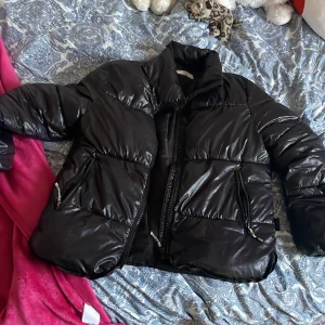 Svart pufferjacka från H&M - Säljer en svart pufferjacka från H&M i storlek 152. Jackan har en glansig yta med dragkedja framtill och två fickor med dragkedjor. Perfekt för kyligare dagar med sin vadderade design men det finns ett litet hål på jackans arm men det syns inte så mycket.