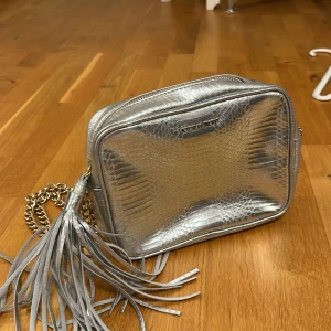 Silverfärgad axelväska från Victoria's Secret - Snygg silverfärgad axelväska från Victoria's Secret med ormskinnsmönster och fransdetalj. Väskan har en kedjerem och dragkedja för säker förvaring. Perfekt för att ge en glittrig touch till din outfit!