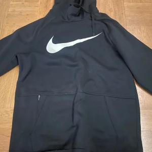 Svart hoodie från Nike - Säljer en svart Nike Therma-Fit hoodie med stor vit logga på bröstet. Tröjan har en stor ficka framtill och en justerbar huva. Perfekt för kyliga dagar. I strl S, knappt använd