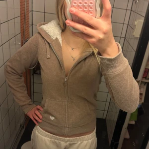 hoodie med foder och hjärtdetalj - Mysig beige hoodie med dragkedja och vit hjärtdetalj på fickan. Insidan är fodrad med fluffigt material för extra värme. Perfekt för kyliga dagar!