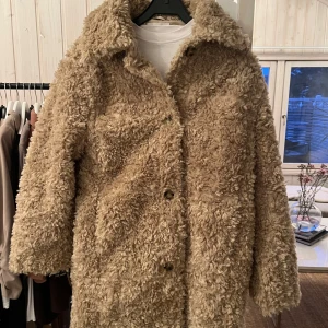 Beige teddyjacka - Mysig beige teddyjacka med knappar framtill. Perfekt för kyliga dagar med sin fluffiga och varma design. Jackan har en klassisk krage och långa ärmar.