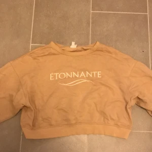 Beige croppad tröja från H&M - Säljer en beige croppad tröja från H&M med texten 'Étonnante' på framsidan. Tröjan har långa ärmar och en avslappnad passform. Perfekt för en casual look. Liten defekt runt halsen