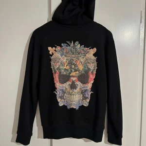 Svart hoodie från Philipp Plein - Snygg svart hoodie från Philipp Plein med en cool dödskalle-design på ryggen. Den har dragkedja framtill och en liten logotyp på bröstet. Perfekt för en stilren och edgy look. Storlek M