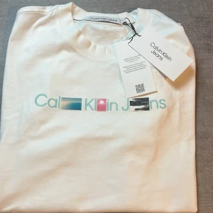 Vit t-shirt från Calvin Klein Jeans - Säljer en stilren vit t-shirt från Calvin Klein med färgglatt tryck på bröstet. Perfekt för en avslappnad look. Modellen är 186 och väg 75 kg. Hör av er vid frågor!
